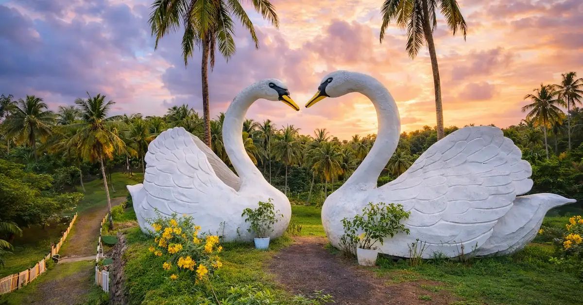 Yellow Garden Port Blair featuring two white swan sculptures forming a heart shape amid lush palm trees and a scenic sunset.