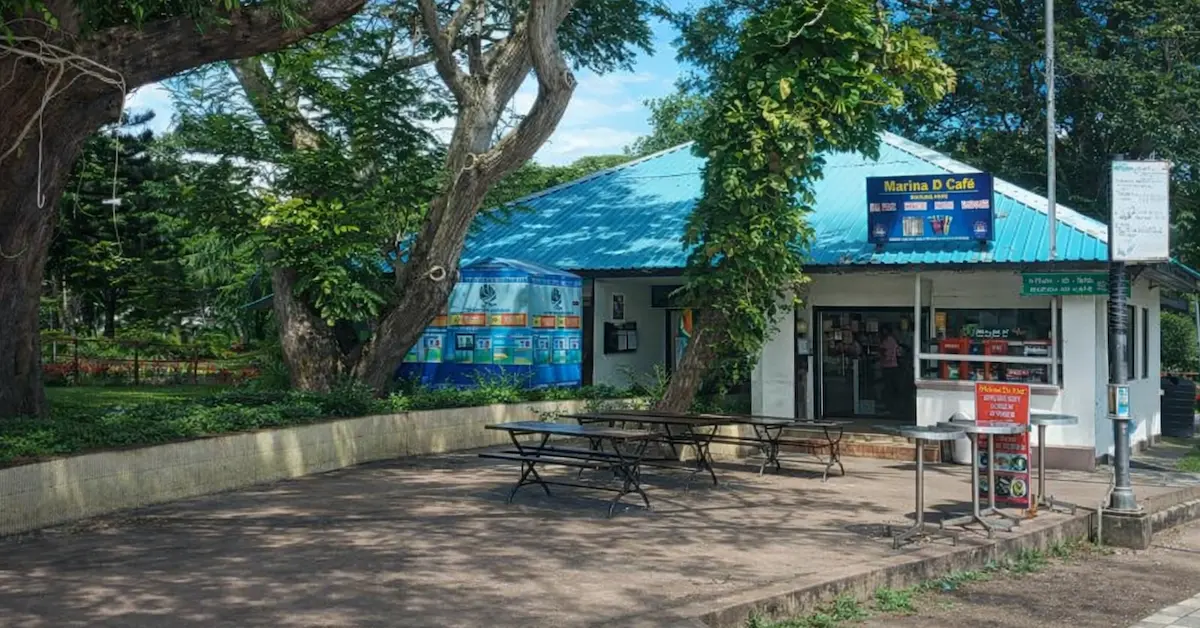A small outdoor café with a blue metal roof surrounded by large trees and greenery, featuring picnic tables on a paved seating area in front, with the café counter visible inside.