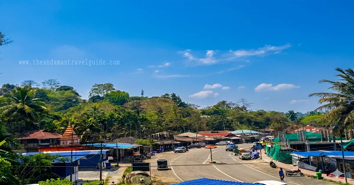 An elevated, wide-angle view of Aerial Bay Bazar in Diglipur, North Andaman. The scene shows a curving asphalt road lined with local shops, houses with colorful blue and red tin roofs, and a small temple structure. The village is nestled against a backdrop of lush green tropical hills under a clear blue sky with scattered clouds.