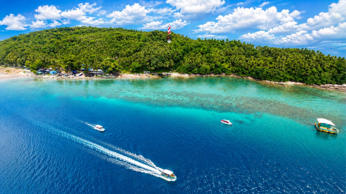 Breathtaking aerial view of North Bay Island Andaman showcasing the lush green coastline, the red and white lighthouse, crystal-clear turquoise coral reefs, and speedboats leaving white water trails in the deep blue sea.